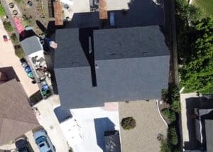 Aerial view of a completed architectural roof with multiple rooflines and a chimney, surrounded by neighboring houses and gre.