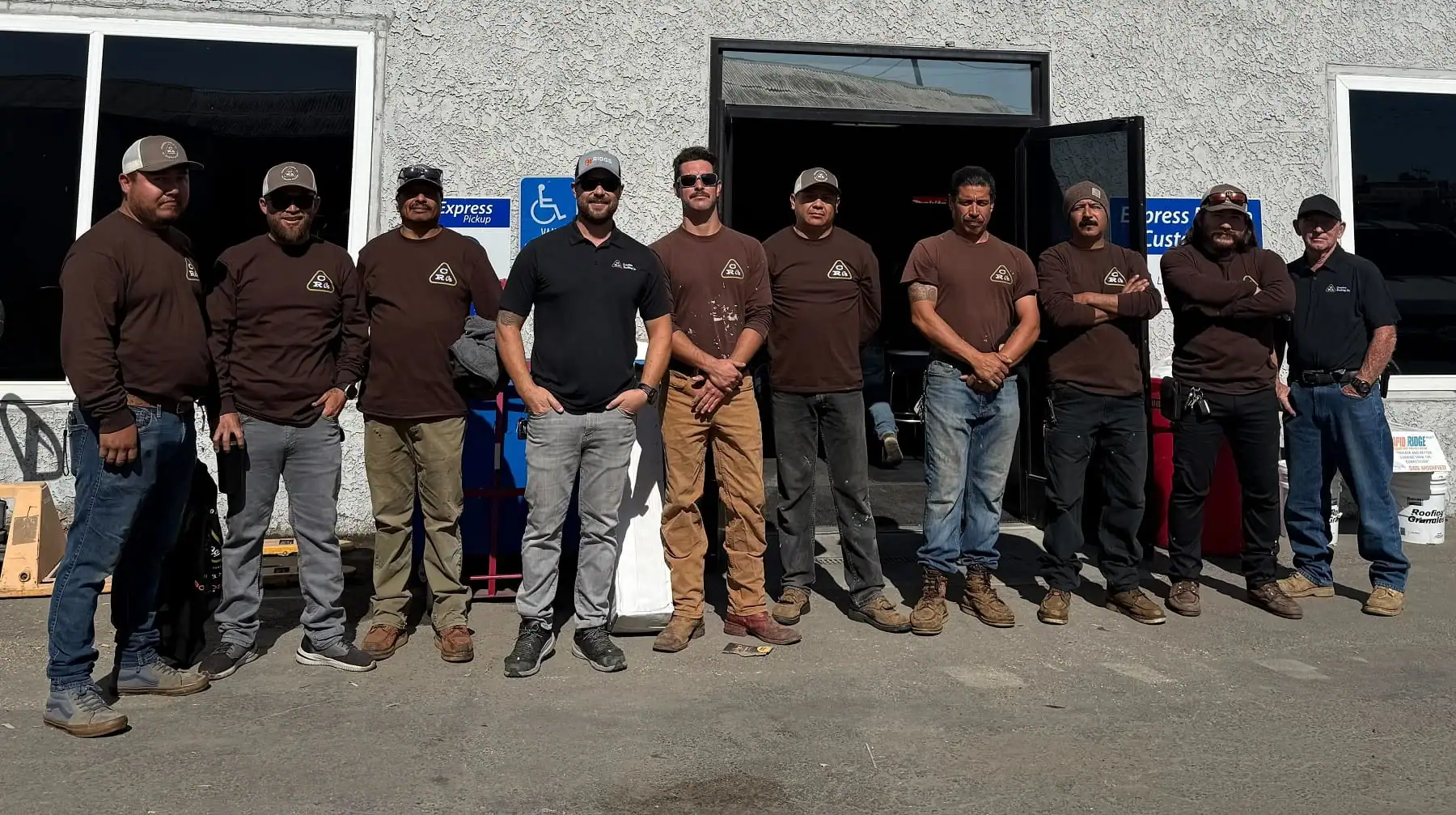 Group of nine roofing professionals standing outside a building with open doors.